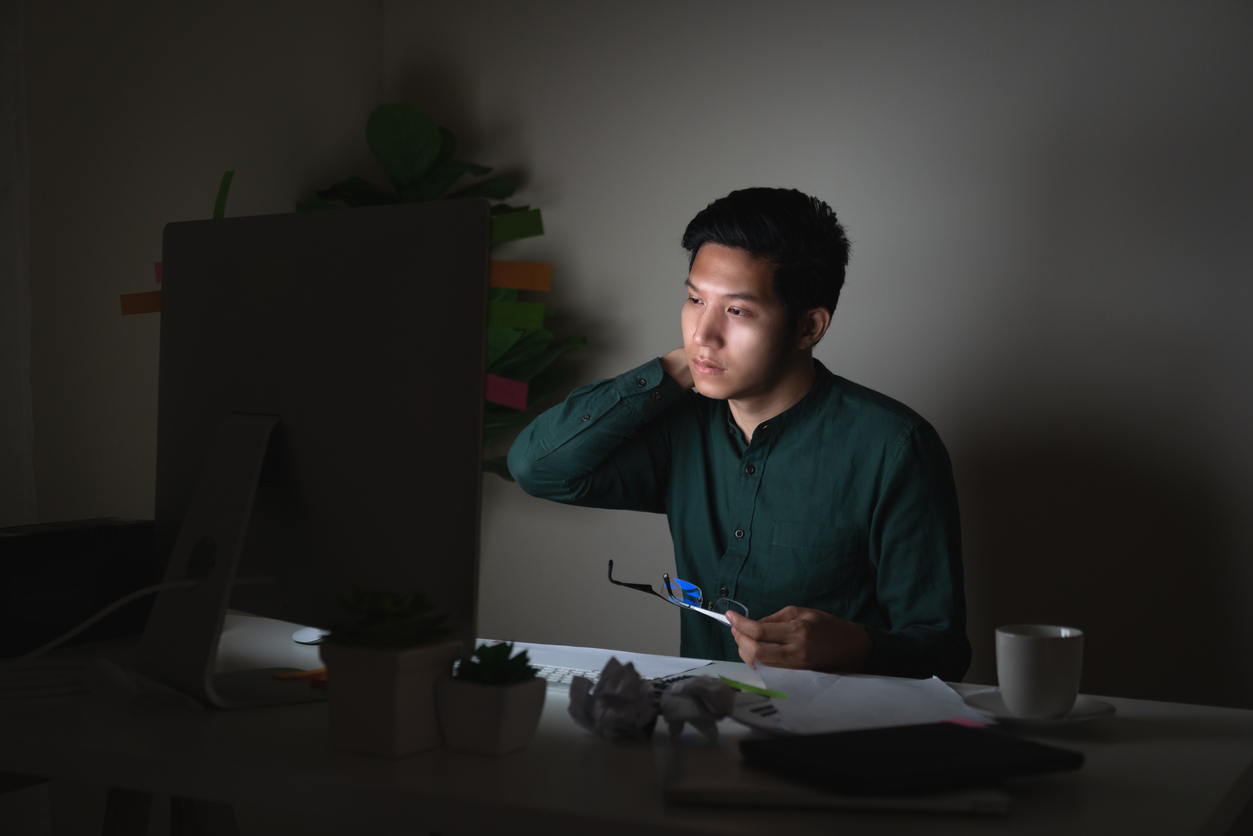 Attractive young asian man working late dark night looking at laptop computer in dark home office desk feeling tired on work load or work hard concept. Overworked male feeling unhappy and sleepless.