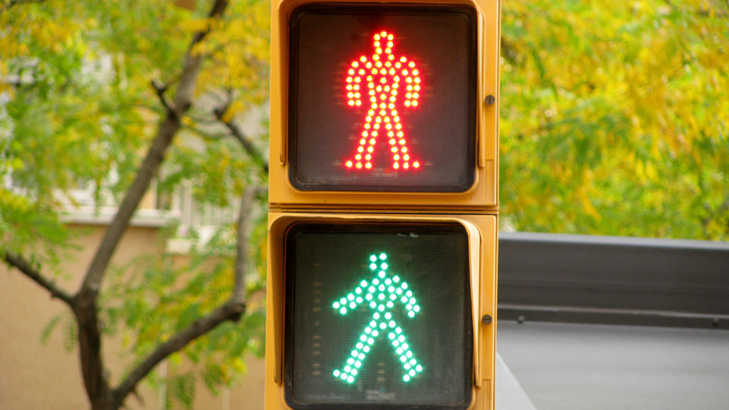 Pedestrian traffic light displaying a red 'stop' figure above a green 'walk' figure, set against a backdrop of trees with yellowing leaves and an urban setting.