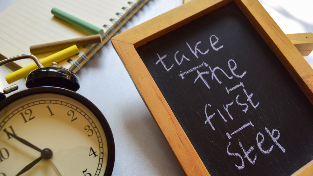 Close-up of a vintage alarm clock showing the time, next to a notepad with colored chalk and a chalkboard sign with the motivational message 'take the first step' written in white chalk.