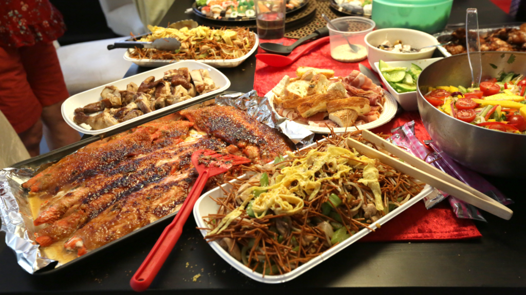 A diverse spread of dishes on a table including a large grilled fish, assorted meats, sliced bread, stir-fried noodles with vegetables, and a vibrant fresh salad in a bowl. Nearby are utensils and a person wearing red is partially visible in the background.