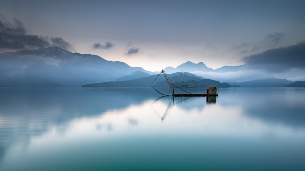"Serenely calm lake reflecting the silhouette of a fishing net structure on a small boat, with misty mountains in the background enveloped by soft morning light and low-hanging clouds.