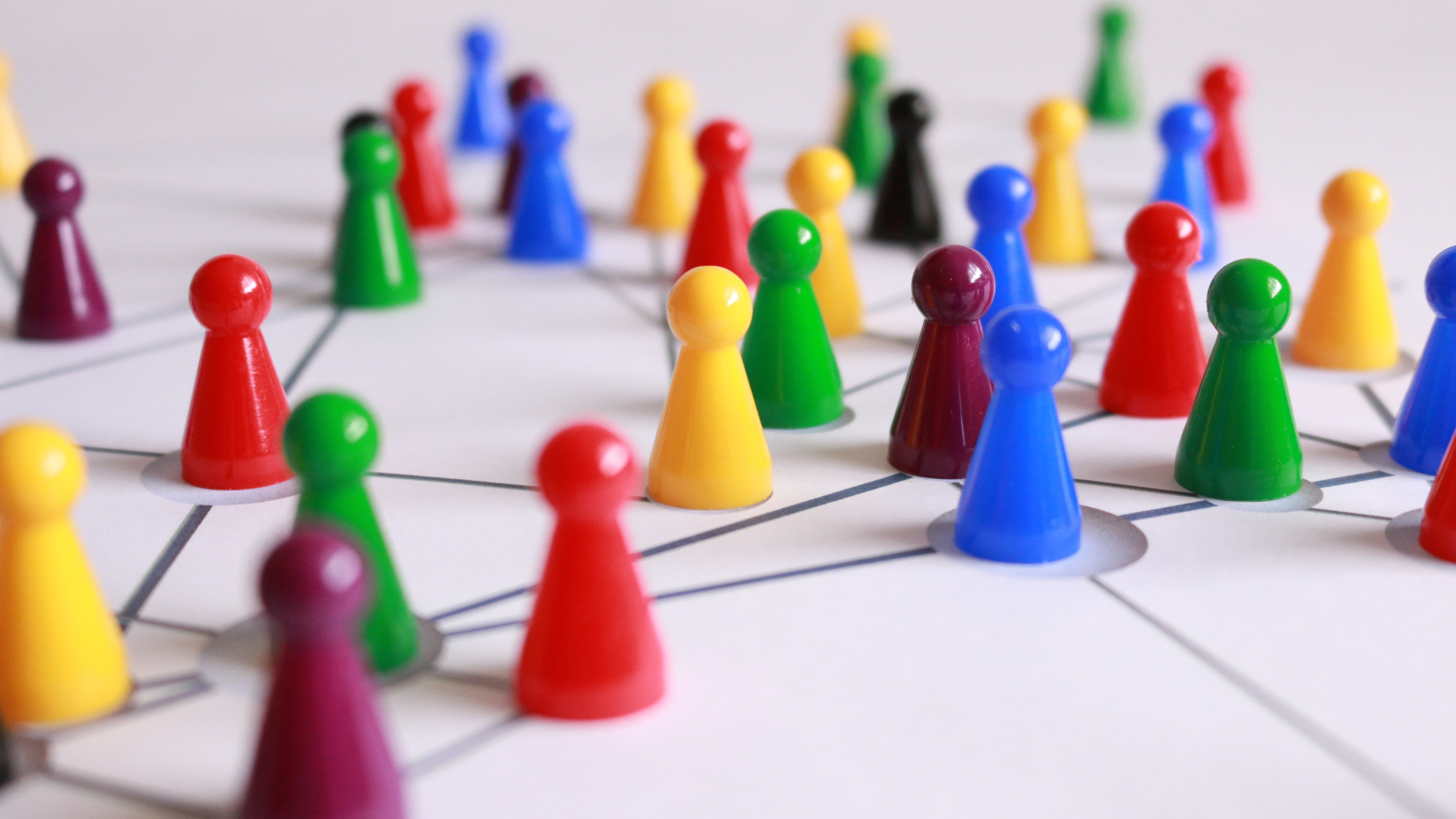 image shows colorful plastic game pieces arranged on a white surface connected by a network of lines. The game pieces are in various colors such as red, yellow, green, blue, purple, and black, symbolizing individuals or groups connected within a broader network or system. The lines between them suggest relationships or connections, illustrating a network structure or concept of teamwork and collaboration.