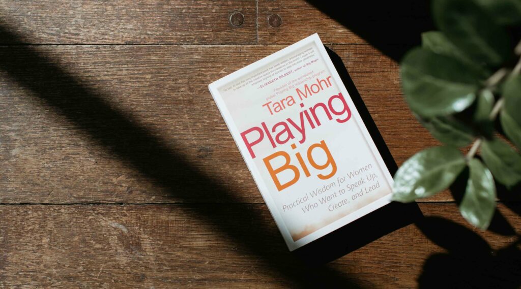 The image shows the book Playing Big by Tara Mohr resting on a wooden surface with a plant partially visible in the lower right corner. Sunlight casts a diagonal shadow across the book, highlighting its cover. The subtitle reads, "Practical Wisdom for Women Who Want to Speak Up, Create, and Lead."