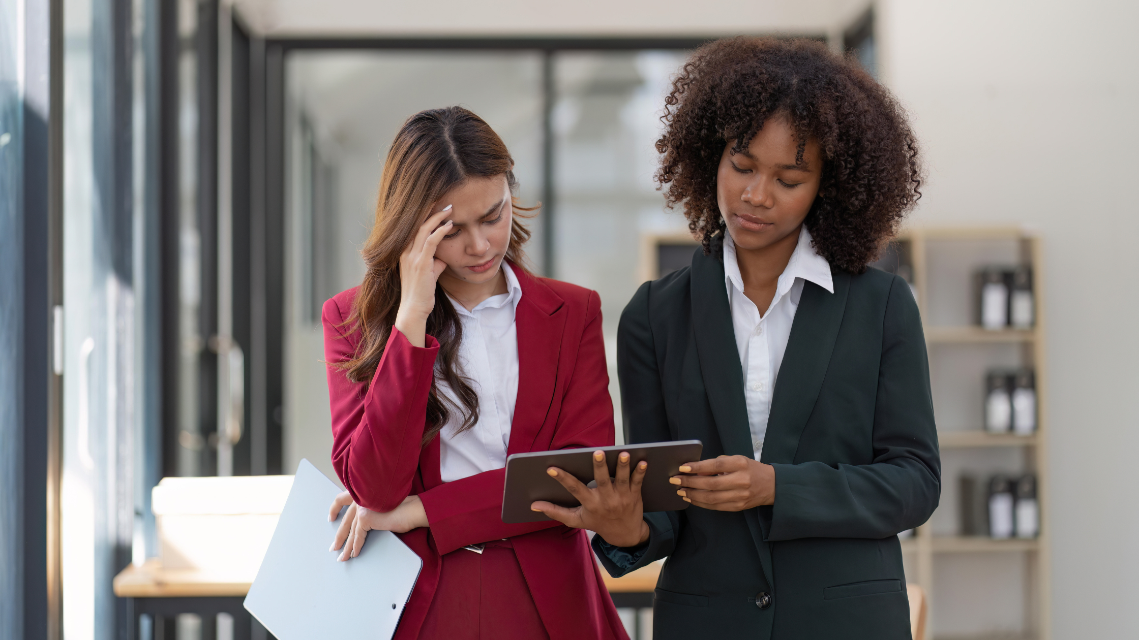 Two women are standing together in a professional office setting. One woman, dressed in a red suit, looks distressed, holding a hand to her head and clutching a tablet. The other woman, wearing a dark green suit, looks calm and focused as she examines the tablet, appearing to offer guidance or support.