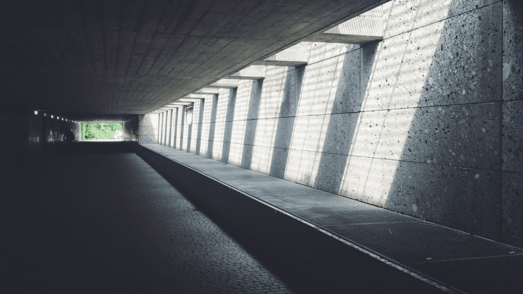 Sunlight streams through a series of large, angled windows along a concrete corridor, casting sharp, parallel shadows on the opposite wall and floor. The passageway leads toward a bright, green exit in the distance, symbolizing structure and clarity illuminated by the light of compassion.