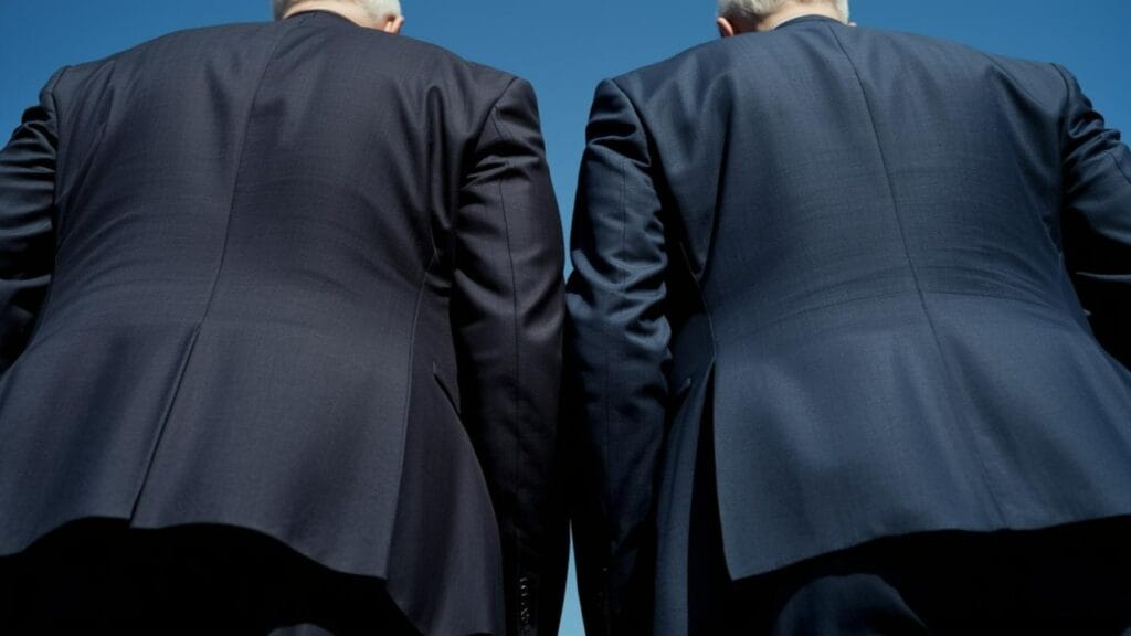 A close-up from behind, two older men stand side-by-side wearing dark navy suit jackets, against a blue sky. Their gray-white hair is visible at the top of the frame. The focus is on the back of their jackets.