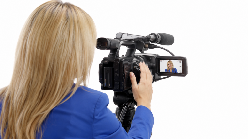 A woman with long blonde hair in a blue blazer films with a professional video camera on a tripod in a bright white studio. Viewed from behind over her shoulder, the camera’s flip-out screen shows her face framed for recording.
